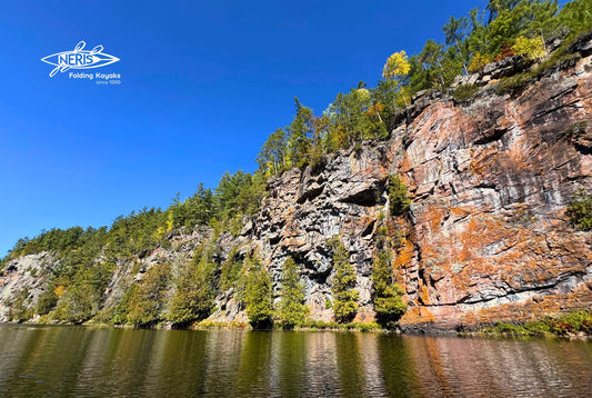 A view of the steep rocky cliffs of Barron Canyon in Algonquin Provincial Park, reflected in the calm waters of the Barron River under a clear blue sky — captured during a kayaking trip with NERIS Folding Kayaks